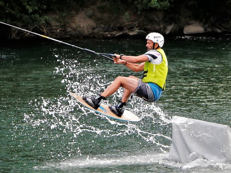 Jeune garçon en wakeboard avec casque et gilet, sautant sur un obstacle blanc sur l'eau.