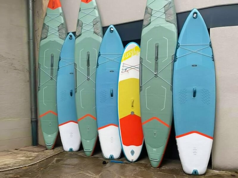 Cinq planches de stand-up paddle de différentes couleurs appuyées contre un mur extérieur.