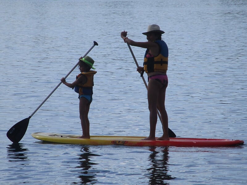 Deux personnes sur un stand-up paddle rouge et jaune pagayant sur un lac calme sous un ciel clair.