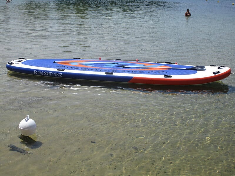 Stand-up paddle bleu, blanc et rouge flottant sur une eau claire près d'une bouée blanche.