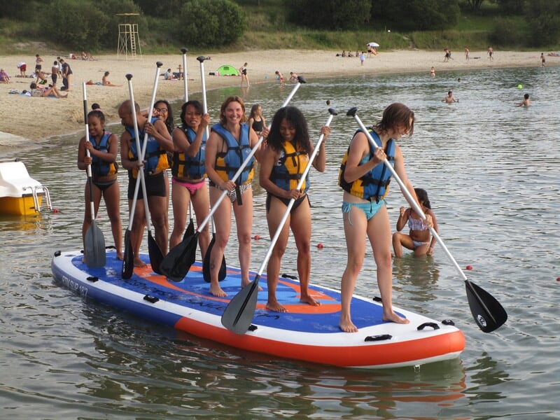 Groupe de jeunes filles debout sur un grand stand-up paddle bleu, blanc et rouge, pagayant au bord d'une plage.