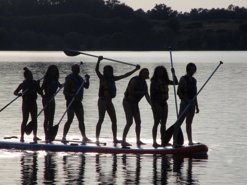 Silhouettes de plusieurs personnes debout sur un grand stand-up paddle au coucher du soleil sur un lac.