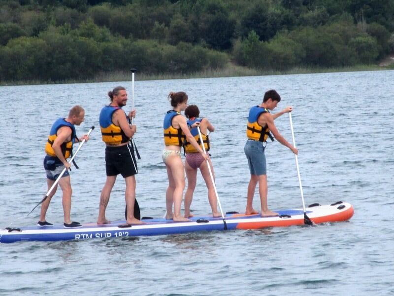 Groupe de personnes debout sur un grand stand-up paddle bleu, blanc et rouge pagayant sur un lac.