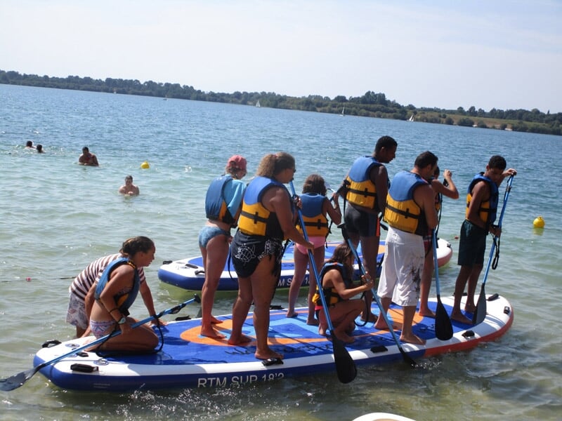Groupe de personnes sur un grand stand-up paddle bleu, blanc et rouge pagayant sur un lac ensoleillé.