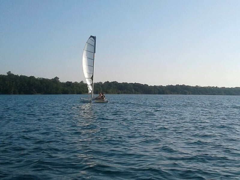 Voilier avec une grande voile blanche naviguant sur un lac calme sous un ciel clair.