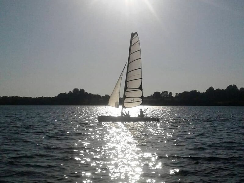 Voilier silhouetté au soleil couchant naviguant sur un lac calme entouré d'arbres.
