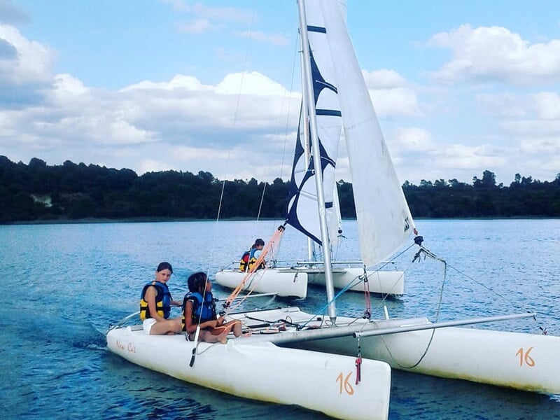 Trois personnes sur un catamaran blanc avec des voiles blanches naviguant sur un lac.