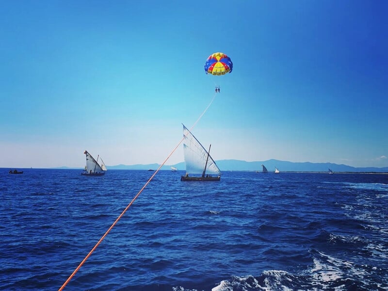 Parachute ascensionnel coloré tiré par un bateau sur une mer bleue avec plusieurs voiliers en arrière-plan.