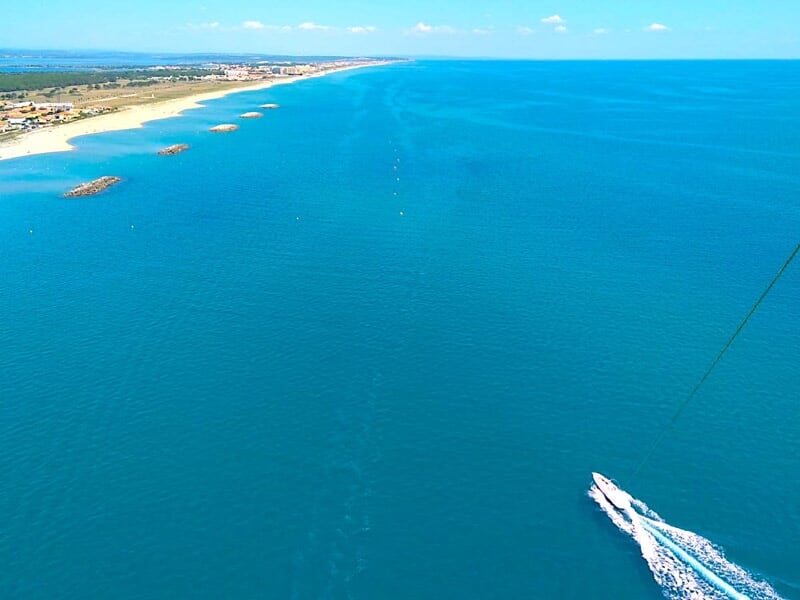 Vue aérienne d'un bateau tirant un parachute ascensionnel au-dessus d'une mer turquoise et d'une plage.
