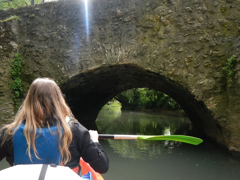 Personne en kayak vue de dos pagayant sous un pont en pierre sur une rivière calme entourée de verdure.