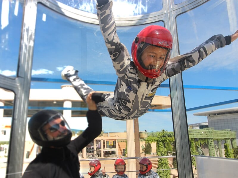 Enfant en combinaison camouflage et casque rouge s'exerce au simulateur de chute libre avec un instructeur à Montpellier.