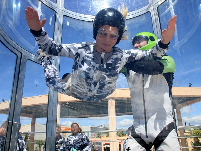 Jeune garçon en combinaison camouflage et casque noir vole dans un simulateur de chute libre avec un moniteur.