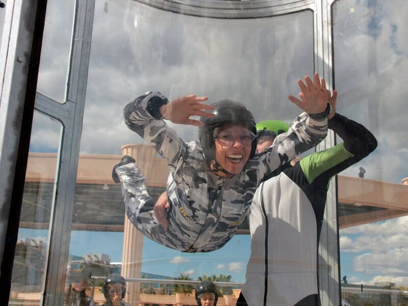 Personne souriante en combinaison camouflage et casque noir s'entraîne au simulateur de chute libre avec un guide.