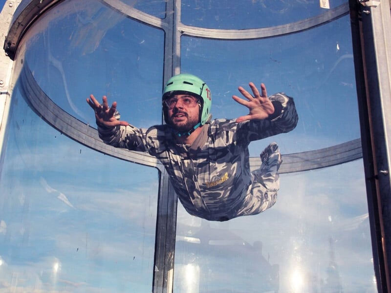 Homme en combinaison camouflage et casque vert pratique la chute libre dans un simulateur transparent à Montpellier.