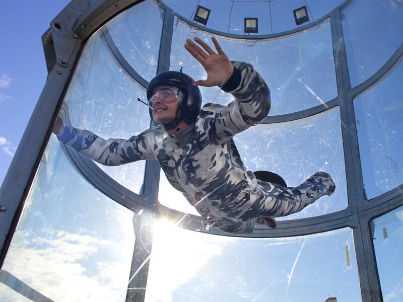 Participant en combinaison camouflage et casque noir vole dans un simulateur de chute libre avec le soleil en arrière-plan.