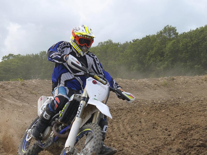 Motocycliste en combinaison bleue et blanche, avec un casque jaune, faisant un virage sur une moto blanche tout-terrain sur un circuit de motocross.