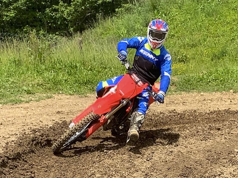 Motocycliste en combinaison bleue et noire, avec un casque bleu et rouge, sur une moto tout-terrain rouge, exécutant un virage serré sur un terrain boueux.