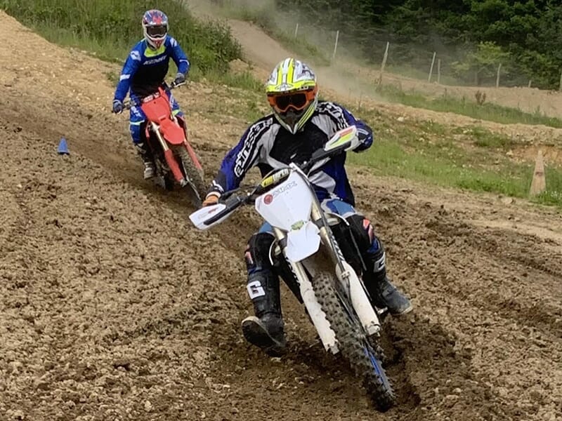 Deux motocyclistes en action sur un terrain de motocross, le premier en combinaison bleue et blanche avec une moto blanche, suivi par un autre en combinaison bleue et noire sur une moto rouge.