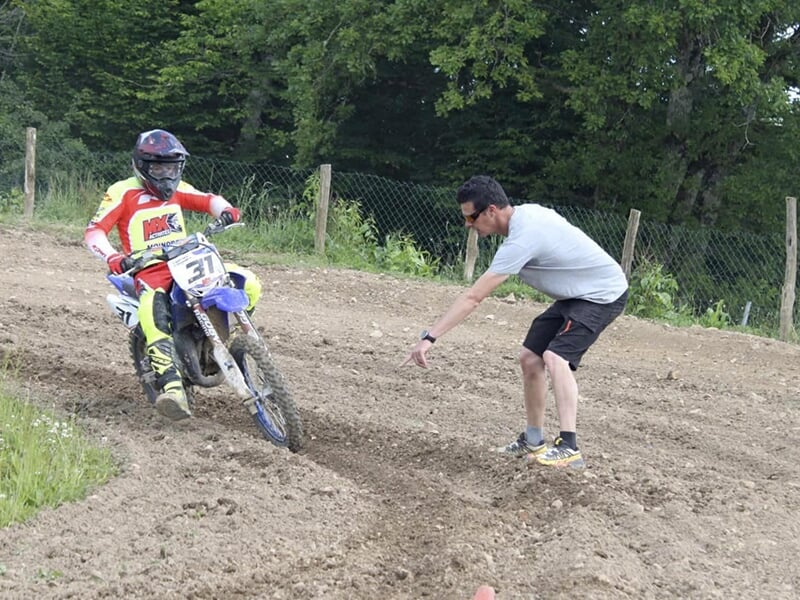 Motocycliste en combinaison rouge et jaune avec un casque noir, écoutant les instructions d'un homme à pied pendant un entraînement sur un terrain de terre.