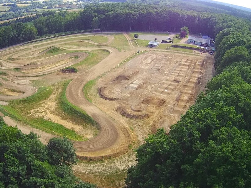 Vue aérienne d'un grand terrain de motocross, montrant plusieurs pistes sinueuses et zones d'obstacles entourées de forêts.