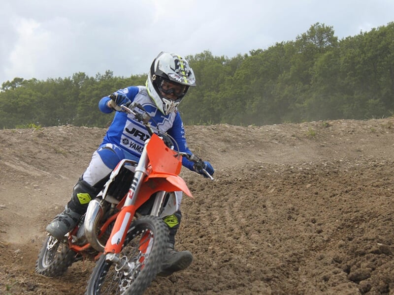 Motocycliste en combinaison bleue et blanche avec un casque noir, conduisant une moto orange sur un terrain de motocross boueux.