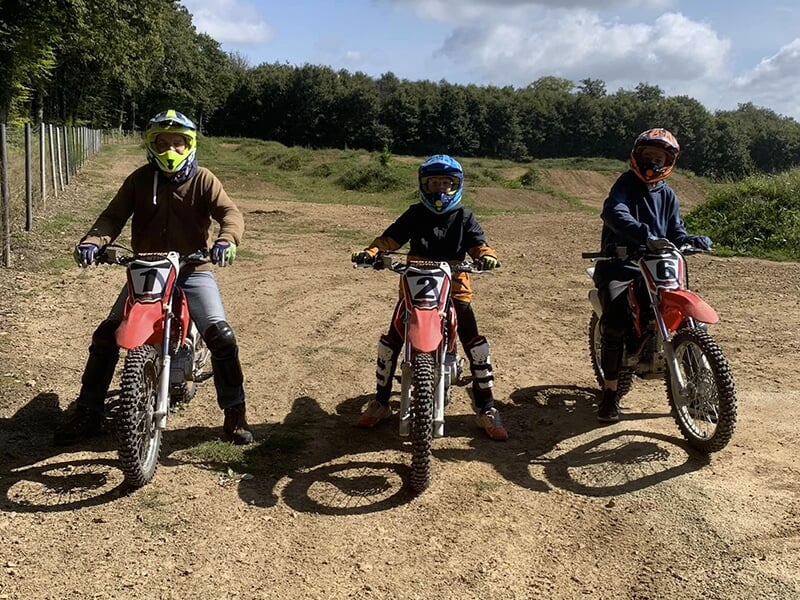 Trois jeunes motocyclistes alignés avec des motos tout-terrain rouges numérotées 1, 2 et 6, prêts à démarrer sur un terrain de motocross.