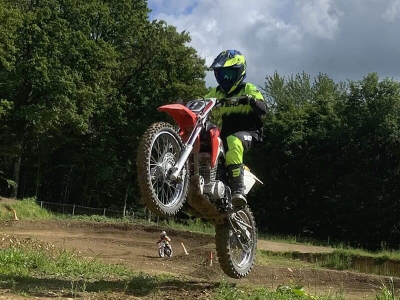 Motocycliste en combinaison verte et noire sautant avec sa moto rouge sur un circuit de motocross, avec un autre motard en arrière-plan.