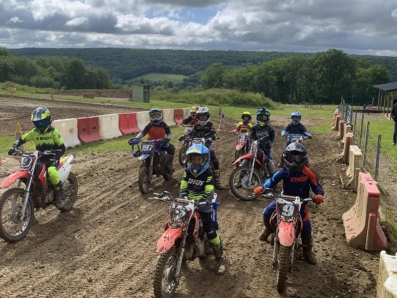 Groupe de jeunes motocyclistes, chacun équipé de casques et sur des motos tout-terrain, se préparant à une course sur une piste boueuse.
