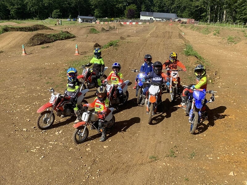Huit motocyclistes en équipement de motocross, posant ensemble sur leurs motos sur un terrain de motocross.