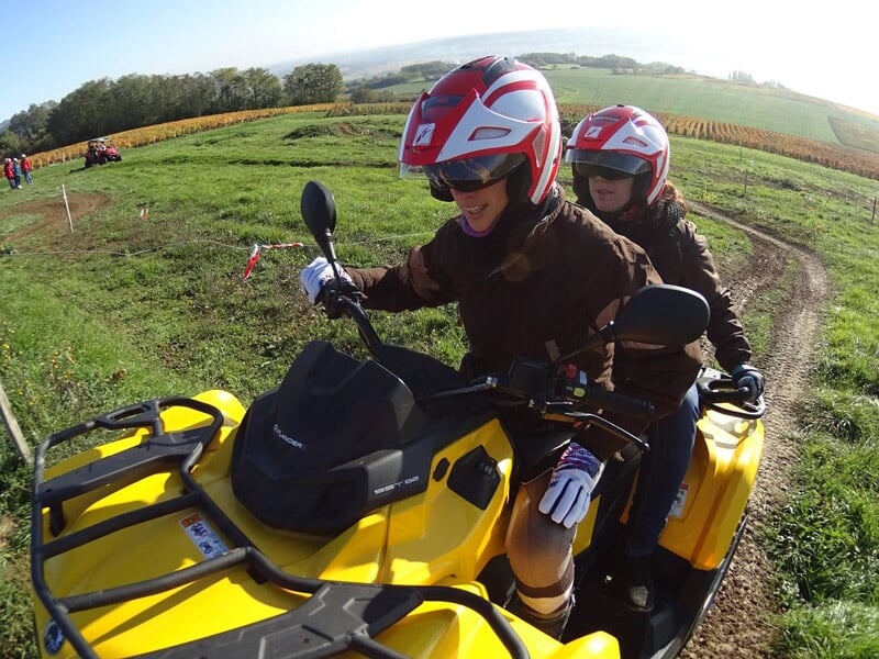 Deux personnes souriantes en quad jaune avec casques rouges, roulant sur un chemin herbeux en campagne.