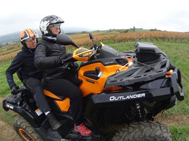 Couple en quad orange tout terrain sur un chemin de terre entouré de vignes sous un ciel nuageux.