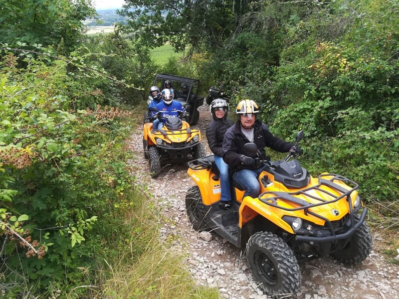Groupe de personnes en quads et buggy tout terrain circulant sur un sentier étroit en forêt dense.