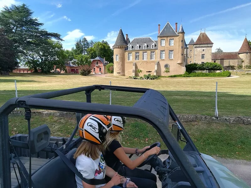 Deux personnes en buggy tout terrain noir avec casques orange devant un château ancien sous un ciel bleu.
