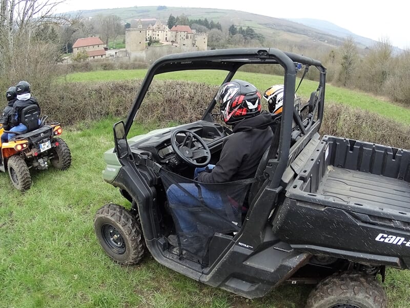 Deux personnes en casque conduisent un quad utilitaire noir sur un chemin herbeux avec un château en arrière-plan.