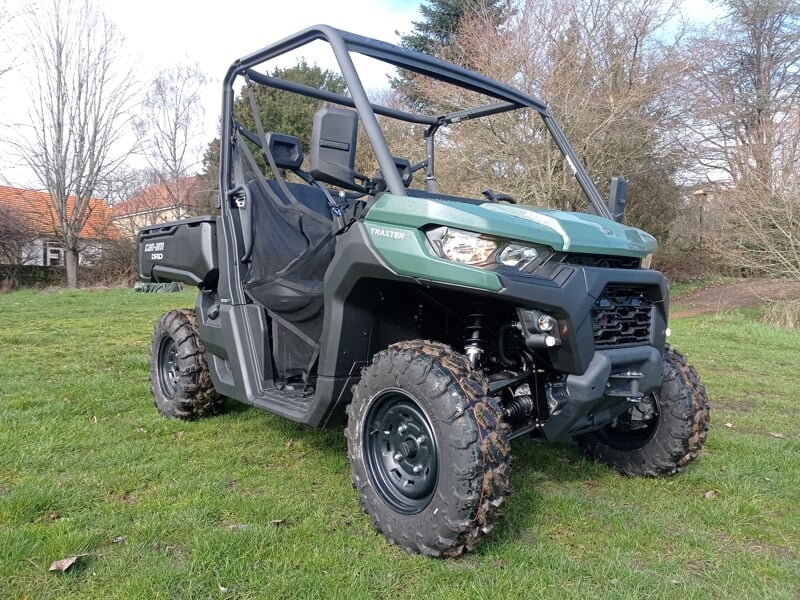 Quad utilitaire vert avec pneus boueux garé sur une pelouse verte, entouré d'arbres et de maisons en arrière-plan.