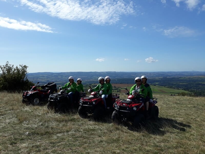 Quads avec des conducteurs en tenue verte et casques, posant sur une colline herbeuse avec un paysage vallonné en arrière-plan.