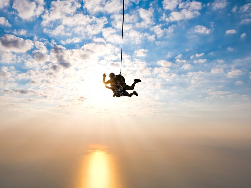 Deux parachutistes en tandem suspendus dans le ciel, ciel nuageux en arrière-plan, reflet du soleil sur l'océan en contrebas