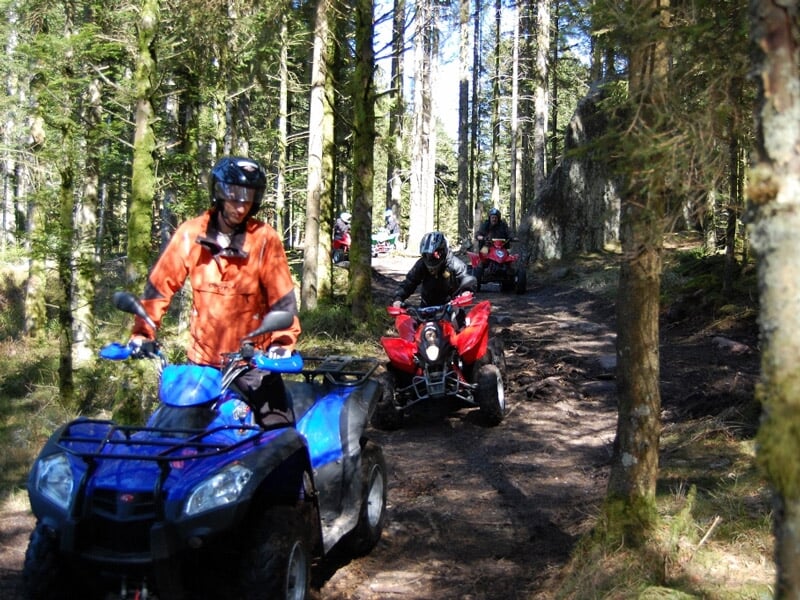 Excursion de quads dans une forêt, pilote en veste orange sur un quad bleu suivi par d'autres quads rouges, arbres verts et sol terreux.