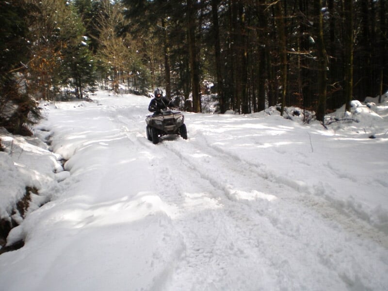 Quad sur chemin enneigé en forêt, conducteur portant un casque noir et une combinaison, arbres entourant le chemin enneigé.