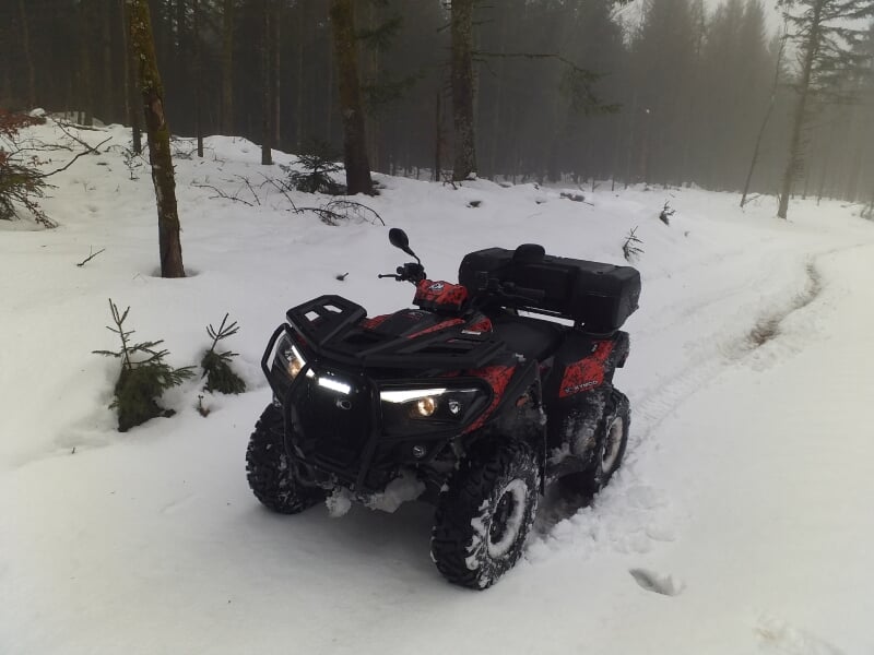 Quad noir et rouge stationné sur un chemin enneigé en forêt, arbres et brouillard en arrière-plan.