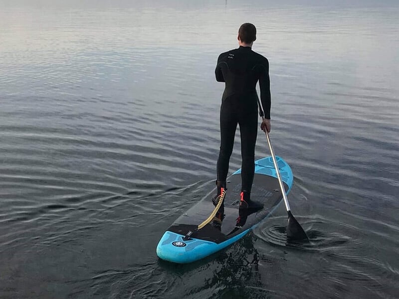 Homme debout sur un paddle bleu pagayant sur une eau calme avec une combinaison noire.