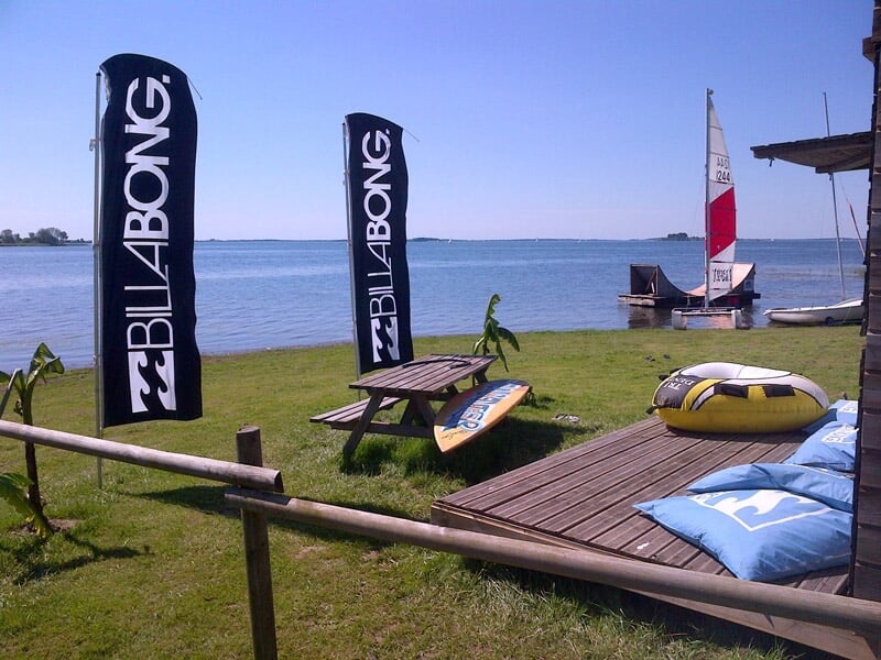 Zone de détente au bord de l'eau avec planche de surf, banc et drapeaux Billabong.