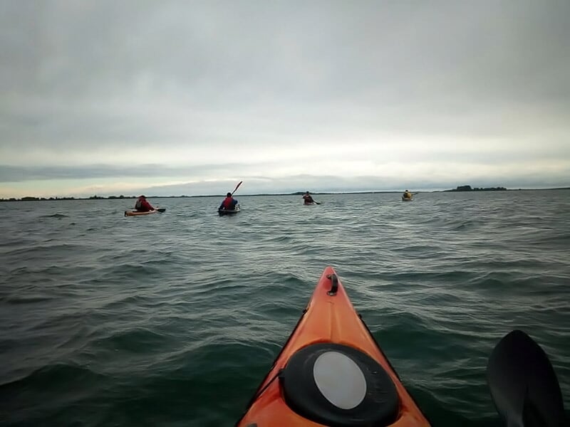 Vue depuis un kayak orange sur une rivière avec plusieurs kayakistes pagayant au loin.