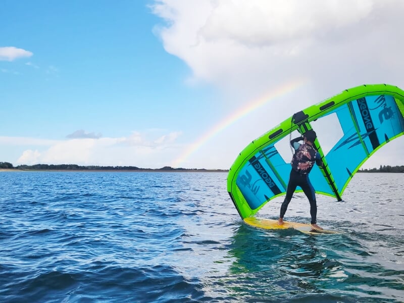 Personne en combinaison noire pratiquant le wingfoil sur un lac sous un ciel bleu.