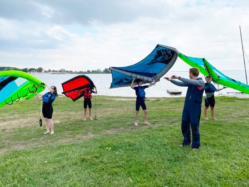 Groupe de personnes apprenant à manipuler des ailes de wingfoil sur une pelouse au bord de l'eau.