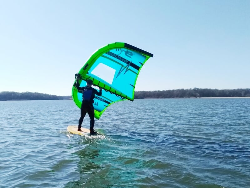 Personne en combinaison noire pratiquant le wingfoil sur un lac avec une aile bleue et verte.