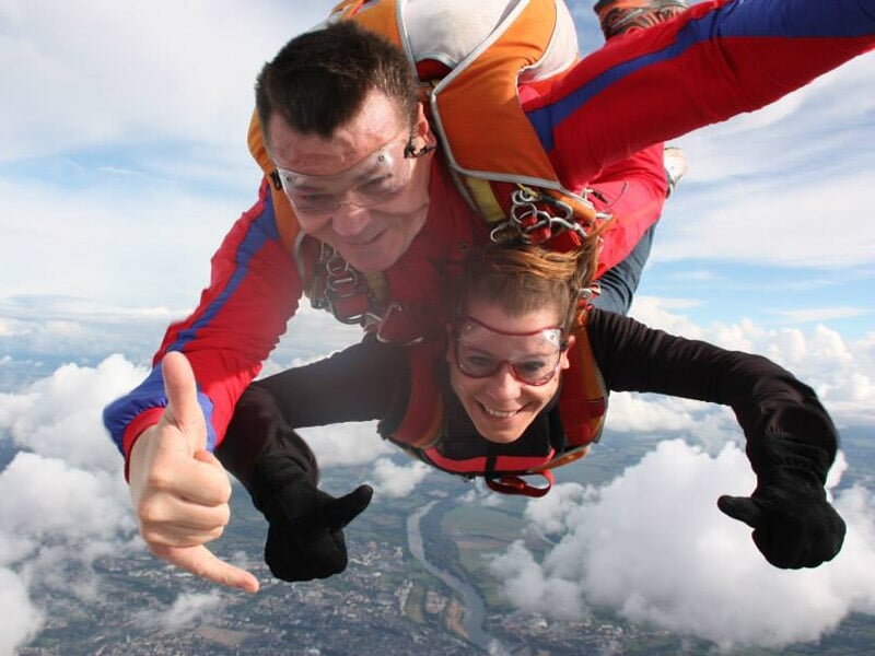 Deux personnes en chute libre lors d'un saut en parachute, ciel bleu et nuages en arrière-plan.