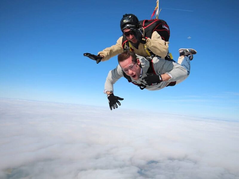 Homme en chute libre encadré par un instructeur en parachute, ciel dégagé et nuages en dessous.