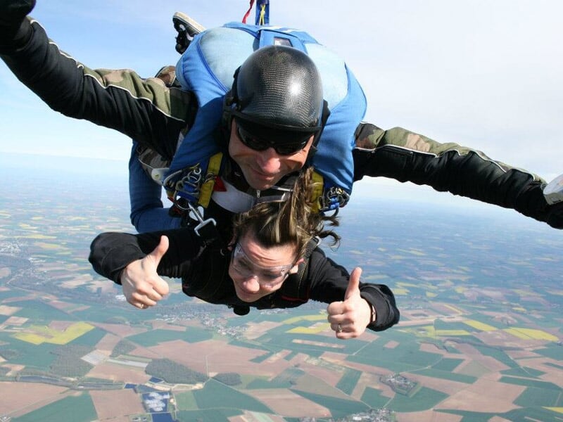 Saut en parachute en tandem, femme souriante avec pouces levés, paysage de champs en arrière-plan.