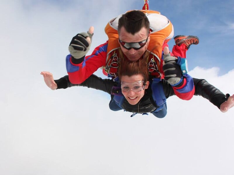 Saut en parachute en tandem, femme souriante avec lunettes de protection, ciel clair en arrière-plan.
